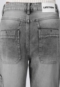 Pantalones de mezclilla grises con un diseño desgastado, dos bolsillos traseros y una etiqueta blanca con "URTRN" en la cinturilla.