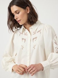 Vrouw met middellang bruin haar, gekleed in een witte blouse met opgeblazen mouwen en bloemvormige uitsneden op de borst en mouwen.