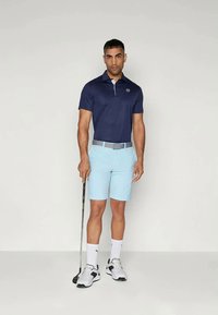 Αθλητική εμφάνιση γκολφ με ναυτικό μπλε polo, ανοιχτό μπλε σορτσάκι, γκρι ζώνη και λευκά παπούτσια. Κρατάει ένα μπαστούνι γκολφ στο αριστερό χέρι.