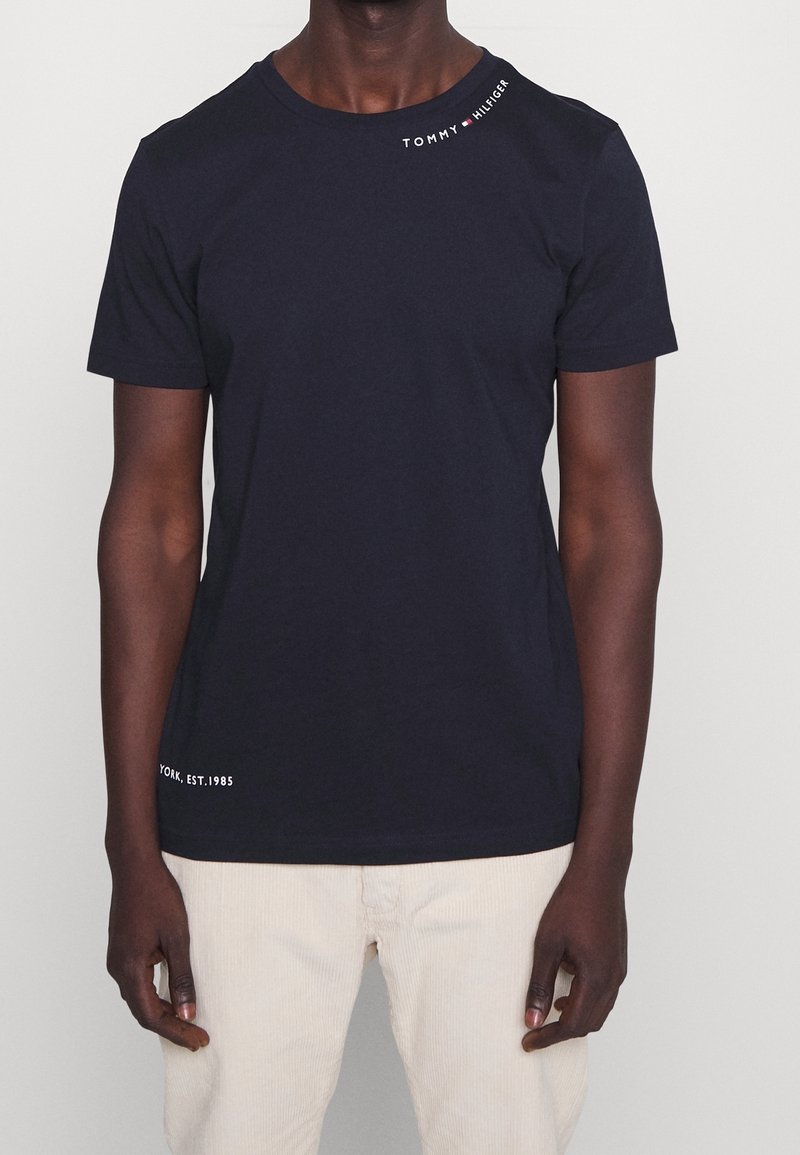 Tmavě modré bavlněné tričko s krátkými rukávy a kulatým výstřihem, s bílým logem "TOMMY HILFIGER" u ramene a nápisem "NEW YORK, EST. 1985" na lemu.