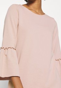 Ljusrosa tröja med klockärmar som har en volangdetalj. Mjuk tyg och rund halsringning. Tätt sittande design, lämplig för lager-på-lager.