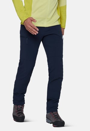 Personne portant une veste jaune, un pantalon bleu marine avec une poche zippée et des chaussures de randonnée grises, debout avec un pied en avant.