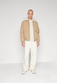 Chaqueta bomber beige con cuello de borrego, dos bolsillos frontales y puños elásticos. Lleva puesta sobre un suéter ligero y combinada con pantalones color blanco roto.