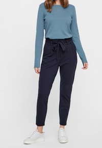 Haut à manches longues bleu clair texturé, associé à un pantalon taille haute bleu marine avec ceinture nouée et poignets resserrés aux chevilles. Porté avec des baskets blanches.