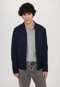 Jūros pajėgų zip-up tipo striukė su pečių siūlėmis, priekinėmis kišenėmis ir lygia tekstūra, dėvima ant dryžuotos marškinėlių ir derinama su pilkais džinsais.