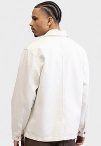 Hvid denimjakke med en lidt oversized pasform, synlige sømme og knaplukninger ved ærmerne, kendetegnet ved en rygskæring og et enkelt design.