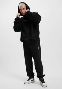 Sort hættetrøje med ribbede manchetter, parret med løstsiddende sorte sweatpants med et hvidt logo. Fodtøjet inkluderer sølv- og sorte sneakers.
