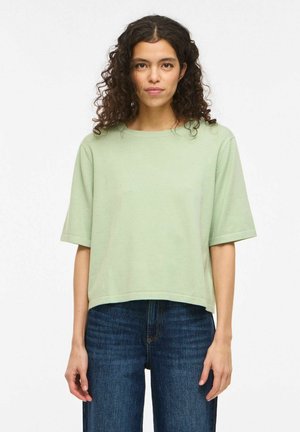 Kvinne med krøllete hår iført en lysegrønn topp med korte ermer og blå jeans, stående mot en enkel hvit bakgrunn.