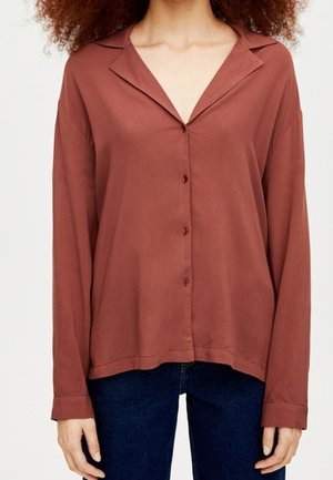 Femme portant une chemise ample couleur rouille à boutons avec un col large et un jean bleu foncé, debout devant un fond uni.
