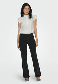 Blusa blanca con volantes y un escote alto, con detalles de botones, combinada con jeans negros de campana. La modelo está de pie con los pies ligeramente separados.