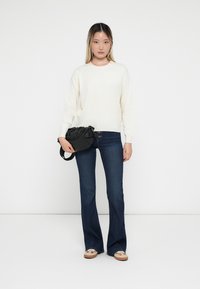 Sweter w kolorze kremowym, ciemne dżinsy dzwony, czarna torba z marszczonym ściągaczem i jasne buty z ozdobnymi akcentami. Prosta, casualowa stylizacja.