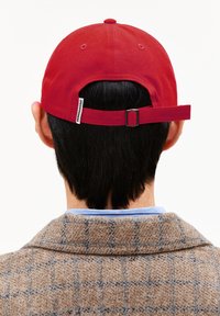 Casquette de baseball rouge avec une sangle en tissu à l'arrière. Présente une petite étiquette sur le côté et un design uni, posée sur une veste en plaid marron texturé.