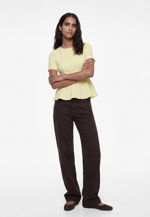 Femme aux longs cheveux foncés, debout les bras croisés, portant un haut jaune texturé à manches courtes, un pantalon marron foncé et des chaussures plates marron.