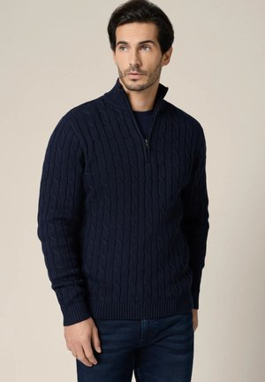 Ciemnogranatowy sweter dziany z wzorem warkocza, wysokim kołnierzem z zamkiem na pół, ściągaczami na mankietach i na dole, zestawiony z niebieskimi dżinsami.