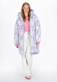 Lang pufferjakke med et pastelmønster af hjerter i blå, pink og gul. Den har en hætte og en frontlynlås, kombineret med lyse jeans.