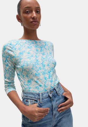 Frau trägt ein weißes und blau gemustertes Langarmshirt, in eine hoch taillierte hellblaue Jeans gesteckt, Hände ruhen nahe den Taschen.