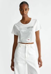 T-shirt bianco corto con maniche corte, caratterizzato da un design grafico a rete di un volto e il testo "UNIQUE" in nero. Abbinato a jeans bianchi a vita alta.