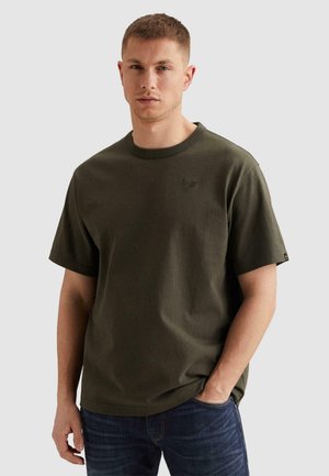 Hombre con camiseta de manga corta de cuello redondo de color verde oscuro y vaqueros azules, de pie con una mano en el bolsillo contra un fondo liso.