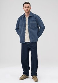 Denimjakke i mørkeblå med to brystlommer og frontknapper, kombineret med beige skjorte og mørkeblå vidbenede jeans. Brune ruskindssko.