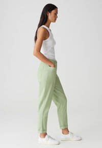 Pantalon vert clair à coupe décontractée, avec deux poches latérales, des ourlets roulés et une fermeture à bouton. Associé à une chemise sans manches blanche.