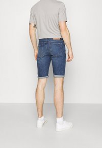 Tummansiniset farkkushortsit, joissa on käännetty helma, yhdistettynä lyhythihaiseen harmaaseen paitaan ja valkoisiin lenkkareihin. Takana näkyvät taskut.