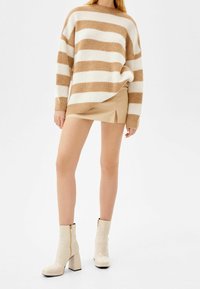 Kobieta w beżowym i białym paskowanym swetrze, beżowej mini spódniczce z bocznym rozcięciem i kremowych botkach na blokowych obcasach na białym tle.
