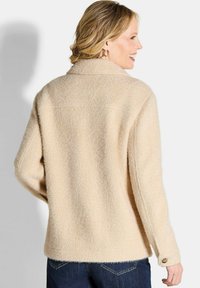 Beige gebreide cardigan met een kraag en knoopmanchetten, met een ontspannen pasvorm en zichtbare naden, gecombineerd met donkere jeans.