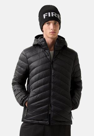 Young man wearing a black puffer jacket with hood and a black beanie with the word "FIRE" in white letters. Hands in jacket pockets.