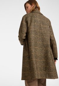 Brązowy i zielony płaszcz w wzór houndstooth o luźnym kroju, z szpiczastym kołnierzem i widocznymi bocznymi kieszeniami. Materiał z mieszanki wełny, w długości średniej.