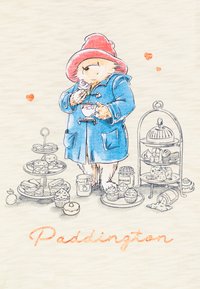 Ilustracija Medvedka Paddingtona prikazuje modro plašč, rdečo klobuk, ki drži sladico, obdan z različnimi pecivi in kozarci, z besedo "Paddington".