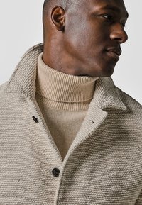 Beige strukturierter Mantel mit einem spitzen Kragen und dunklem Knopf, über einem hellbeigen gerippten Rollkragenpullover getragen.