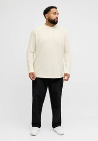 Langermet kremfarget henley-skjorte med tre knapper, kombinert med svarte bukser og hvite joggesko; jevn tekstur og avslappet passform.