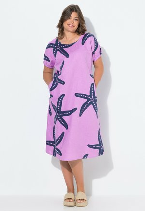 Robe violette à coupe ample, ornée d'un motif d'étoile de mer en bleu foncé. Manches courtes et poches latérales. Portée avec des sandales à brides beiges.