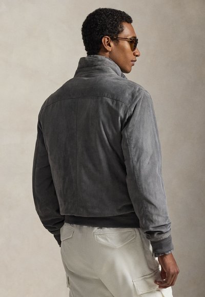 Γκρι, απαλό δερμάτινο μπουφάν τύπου μπόμερ με γιακά, ριμπ μανσέτες και τελείωμα· μοντέλο φοράει ανοιχτόχρωμο παντελόνι τύπου κάργκο, εμφανιζόμενο από πίσω.