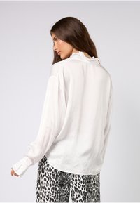 Hvit, luftig bluse med lange ermer og volangdetalj på halsutringning og mansjetter; kombinert med bukser med svart-hvitt leopardmønster.