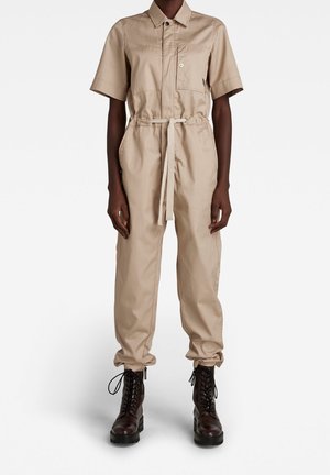 Persoon die een beige jumpsuit met korte mouwen draagt, met een borstzak aan de voorkant en een ceintuur in de taille, gecombineerd met donkerbruine veterlaarzen.