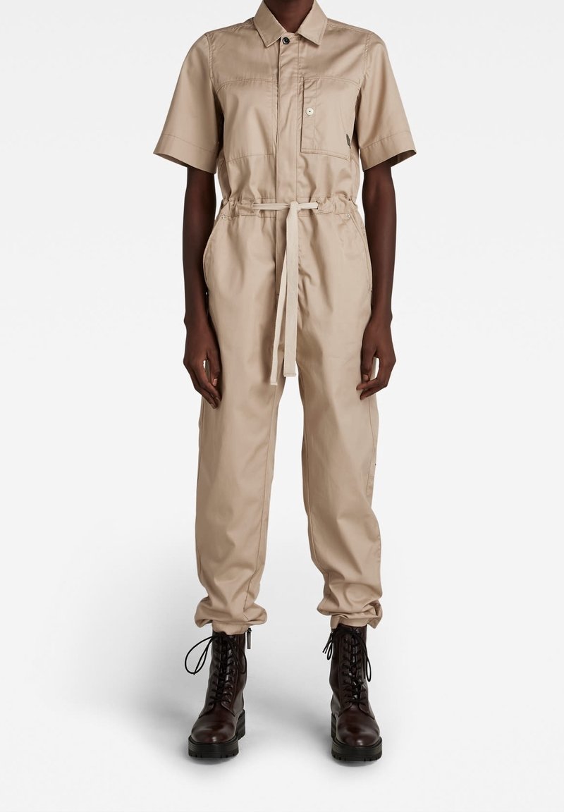 Personne portant une combinaison beige à manches courtes avec une poche poitrine à l'avant et une ceinture nouée à la taille, associée à des bottes lacées marron foncé.