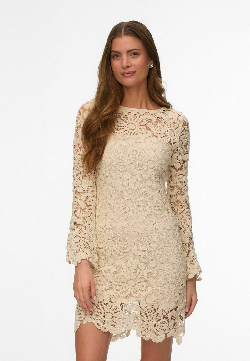 Femme aux longs cheveux bruns portant une robe beige à dentelle florale à manches longues, debout devant un fond blanc uni.