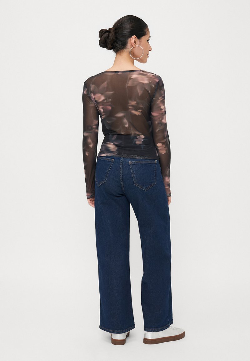 Durchsichtige Langarmbluse mit Blumenmuster, kombiniert mit hoch taillierten, weiten Jeans. Das Outfit wird ergänzt durch silberne Slipper und große Creolen.