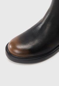 Botte en cuir noir avec un bout arrondi présentant un dégradé de brun, et une semelle noire épaisse sur fond blanc.