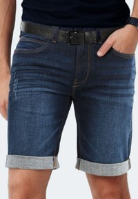 Dunkelblaue Jeansshorts mit Umschlagsaum, ausgestattet mit gelben Nähten und einer Fronttasche. Getragen mit einem schwarzen Ledergürtel.
