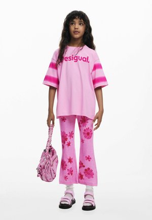 Tricou roz supradimensionat cu textul „desigual.”, pantaloni evazați cu imprimeu floral și geantă roz. Încălțăminte formată din sandale roz și șosete albe.