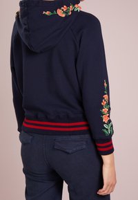 Marinblå sweatshirt med blommönsterbroderi på huva och ärmar, med ribbade muddar och midja i röd- och vitrandigt mönster, mjuk textur.
