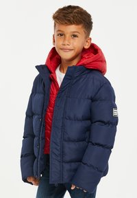 Marineblå pufferjakke lagdelt over en rød quilted hoodie. Har ribbede manchetter, lynlåslukning og logo-patch på venstre ærme.