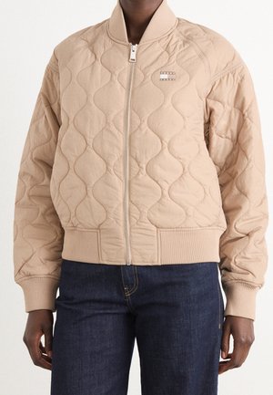 Beige doorgestikte bomberjack met voorrits, geribde boorden en zoom, gedragen met donkerblauwe spijkerbroek.