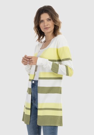 Frau mit schulterlangem Haar, die eine weiß-gelb-grün gestreifte Strickjacke über einem weißen Top und blauen Jeans trägt, stehend vor einem einfarbigen Hintergrund.