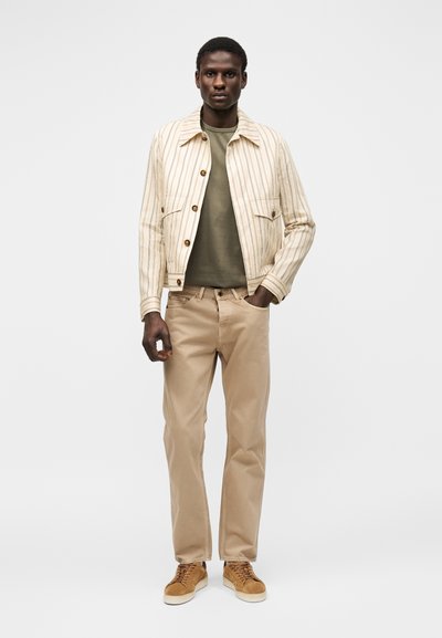 Homme debout portant une veste beige rayée, une chemise vert olive, un pantalon marron clair et des baskets beige sur un fond uni.