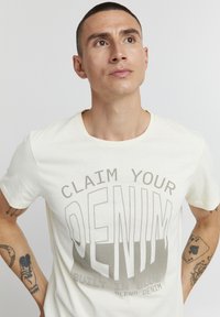 Camiseta de algodón color crema con un destacado texto gris que dice "RECLAMA TU DENIM" y un efecto de degradado. Cuello redondo y mangas cortas.