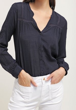 Frau trägt eine marineblaue langärmelige Bluse mit Knöpfen und weiße Hose, Hände in den Hosentaschen.