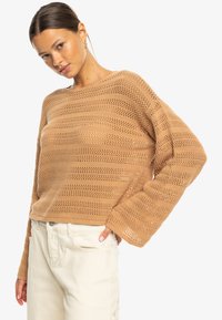 Knit sweater in light brown with open weave design. Features long sleeves and a relaxed fit. Paired with light-colored pants.